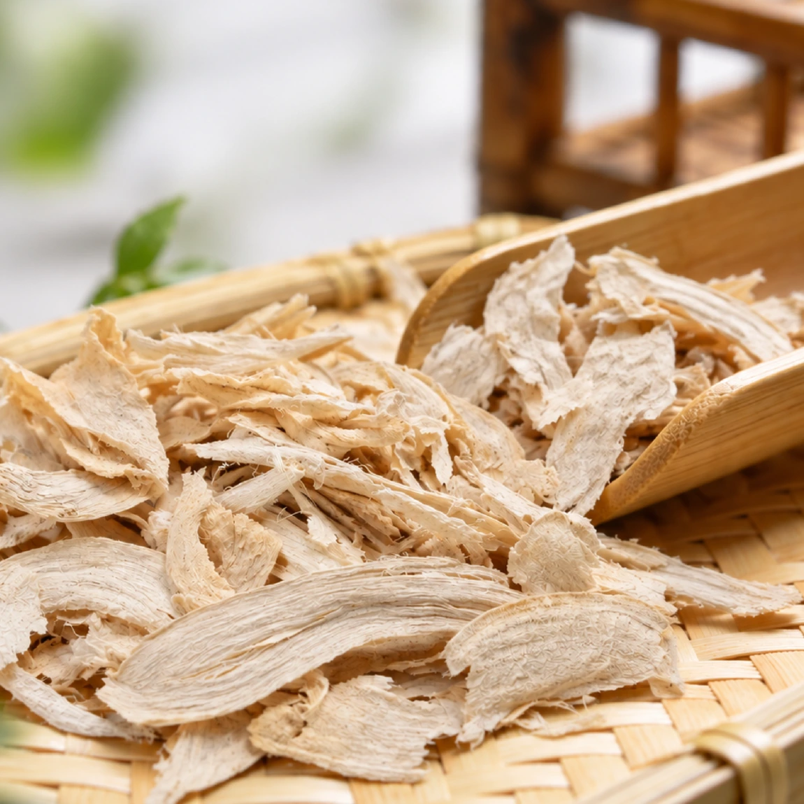 Dried White Kwao Krua (Pueraria mirifica) root slices on a woven bamboo tray, with a bamboo scoop holding more slices.
