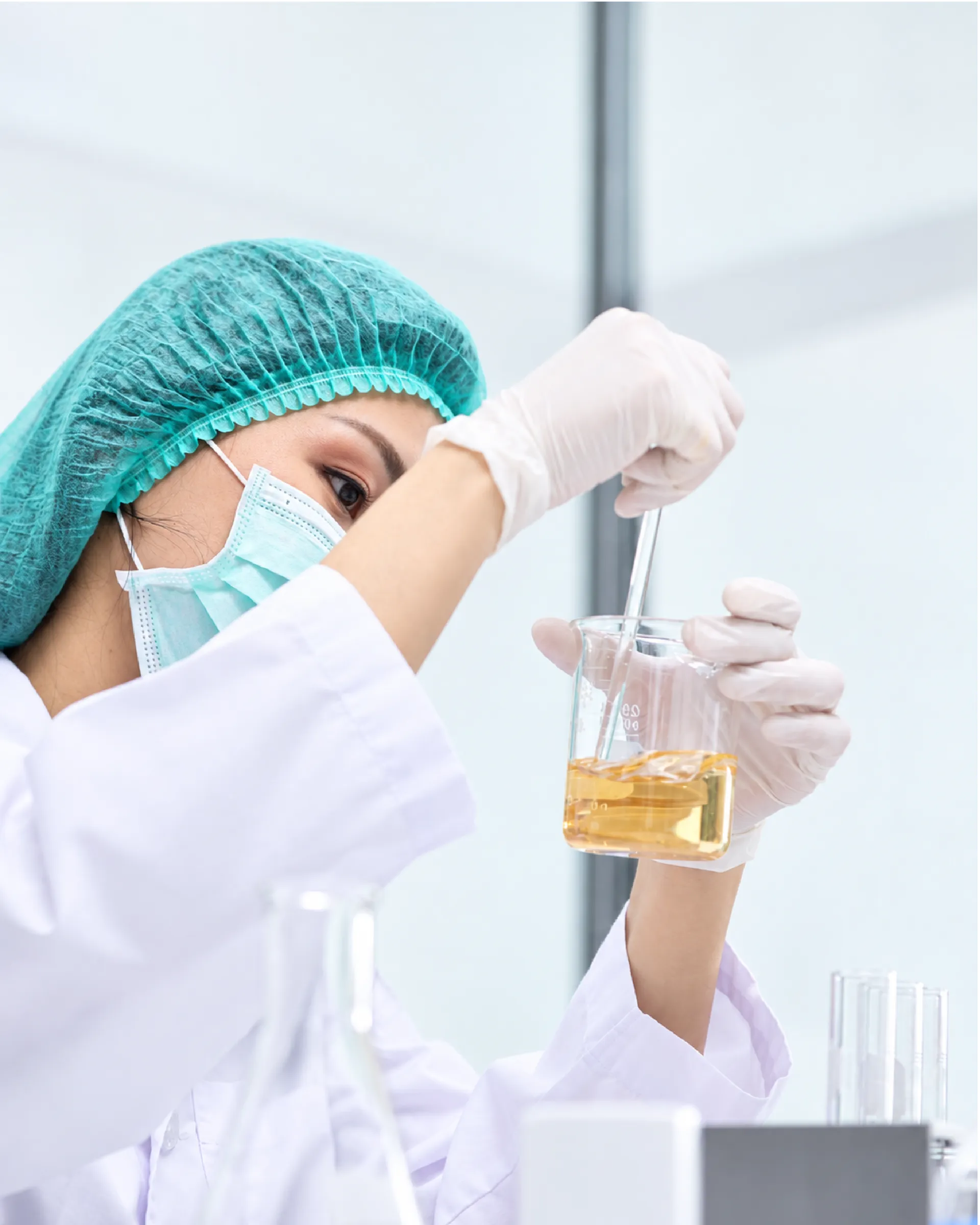 Laboratory technician in a hairnet, mask and gloves stirring an amber liquid in a glass beaker